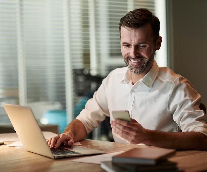 Homme souriant en regardant son téléphone portable assis à un bureau devant son pc portable
